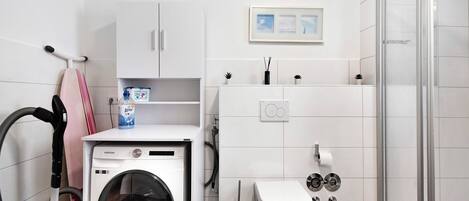 Ein kleines Hotelbad mit Waschmaschine, Toilette, Dusche und einer Ablage für Pflegeprodukte.
