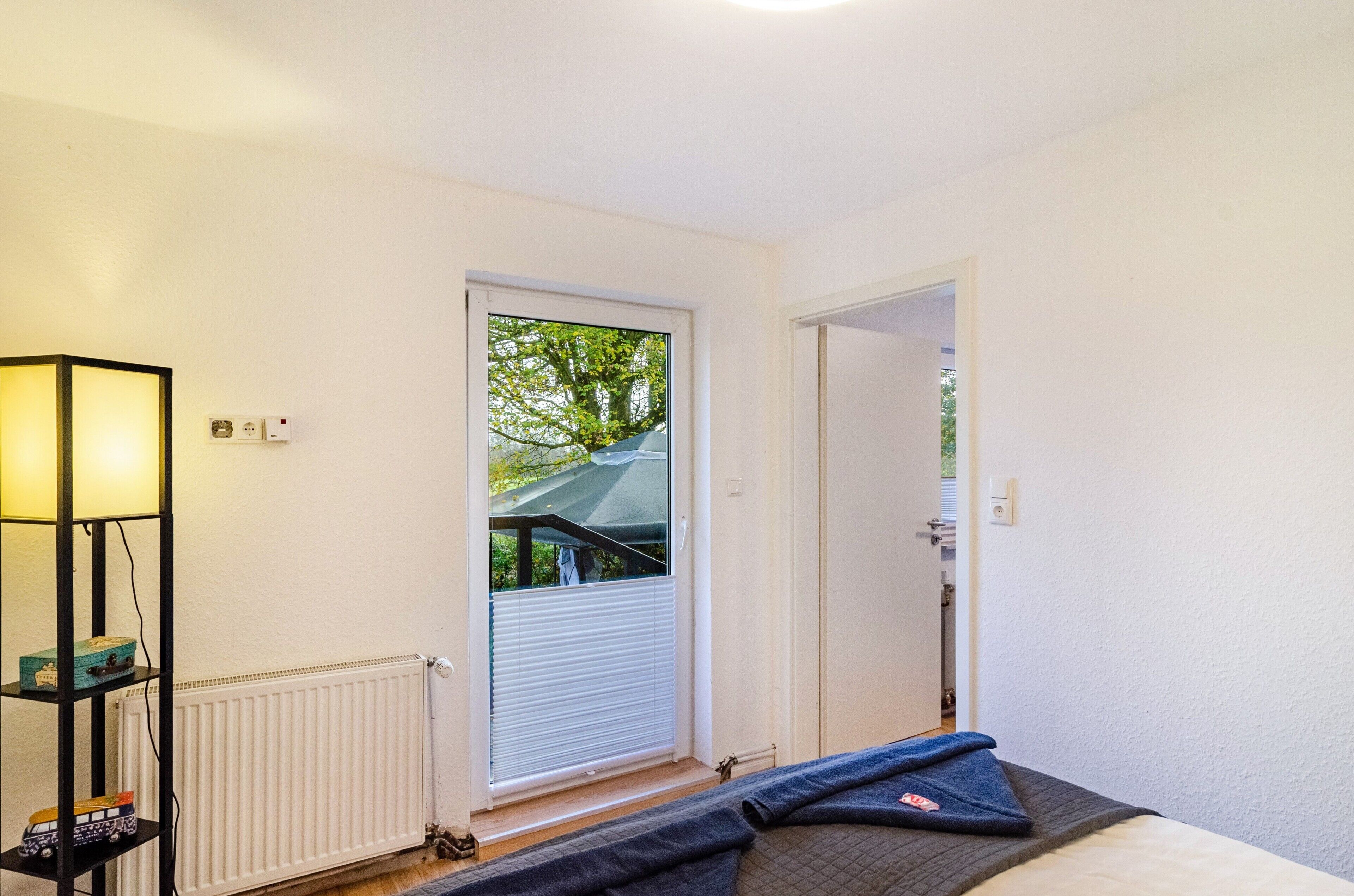 Une pièce comprenant un lit, un radiateur, une étagère avec divers objets et une porte donnant sur une autre pièce.