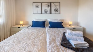 Una cama doble con ropa de cama blanca, dos almohadas azules, una mesita de noche con una lámpara y tres cuadros enmarcados en la pared.