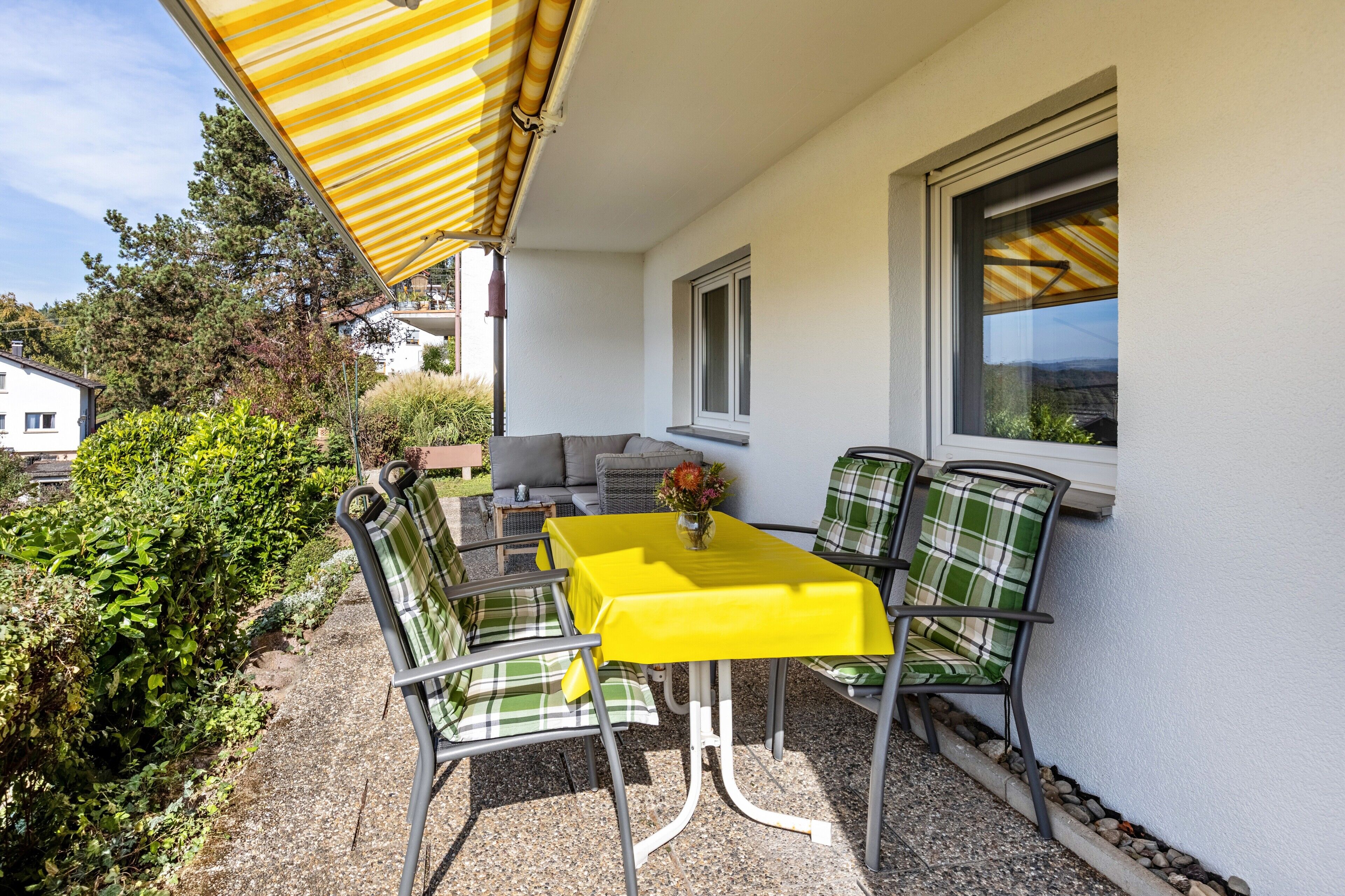 Une terrasse extérieure avec une table jaune et des chaises à carreaux verts, un canapé et une fenêtre donnant sur la verdure.