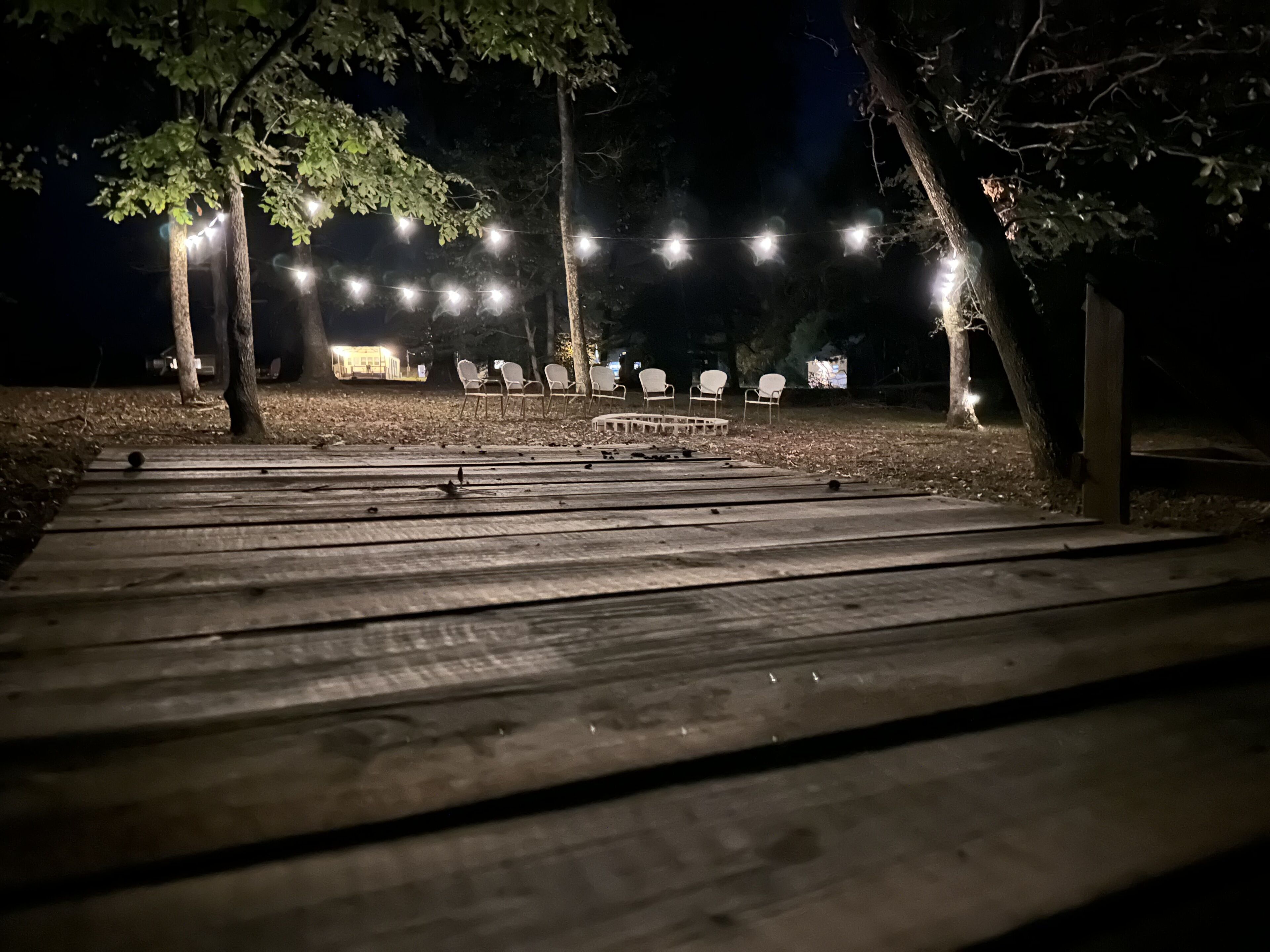 Una terraza de madera que conduce a un área de descanso con sillas blancas, bajo luces colgantes por la noche.
