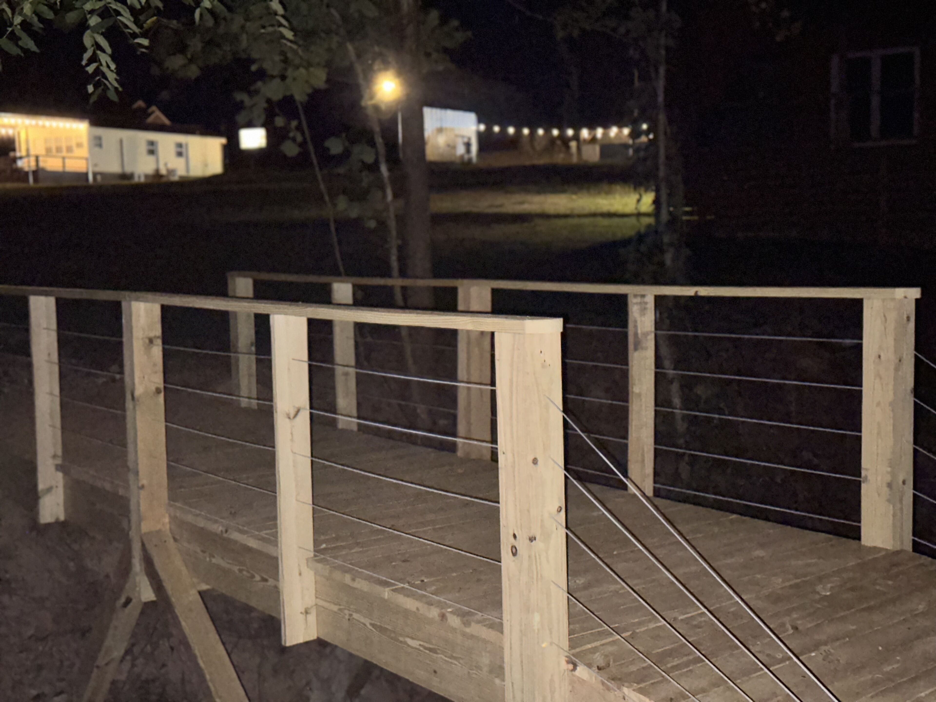 A wooden bridge with metal railings, surrounded by trees and a building with lights in the background.