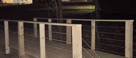 A wooden bridge with metal railings, surrounded by trees and a building with lights in the background.