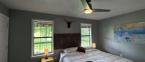 A bedroom with a bed, a ceiling fan, two windows with blinds, a nightstand, a lamp, a deer head mounted on the wall, and a painting.