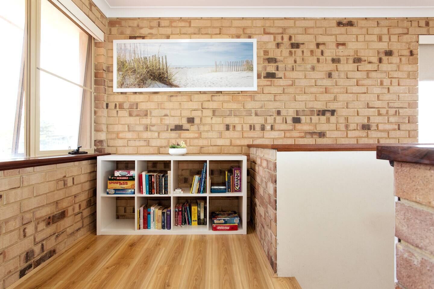 A room with a brick wall, a bookshelf filled with books, a window, and a painting of a beach scene.