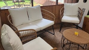 Een overdekt terras met rieten meubilair, waaronder een bank en stoelen, en een ronde tafel met een glas sinaasappelsap.