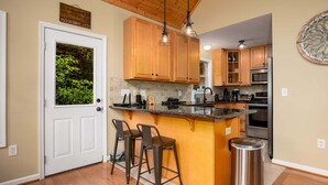 A kitchen with wooden cabinets, a central island with stools, a stainless steel trash can, and a door with a view of greenery. - Moonshine Mountain | Hot tub & fire pit | Bryce Resort (Basye)