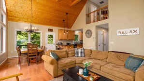 A spacious living room with a large brown sectional sofa, a dining area with a wooden table and chairs, a kitchen in the background, and a 'RELAX' sign on the wall. - Moonshine Mountain | Hot tub & fire pit | Bryce Resort (Basye)