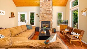 A living room with a stone fireplace, wooden ceiling, and large windows. - Moonshine Mountain | Hot tub & fire pit | Bryce Resort (Basye)