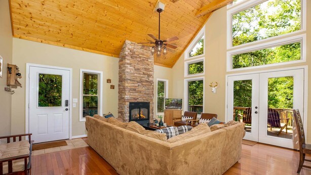 A spacious living room with a stone fireplace, wooden ceiling, and large windows. - Moonshine Mountain | Hot tub & fire pit | Bryce Resort (Basye)