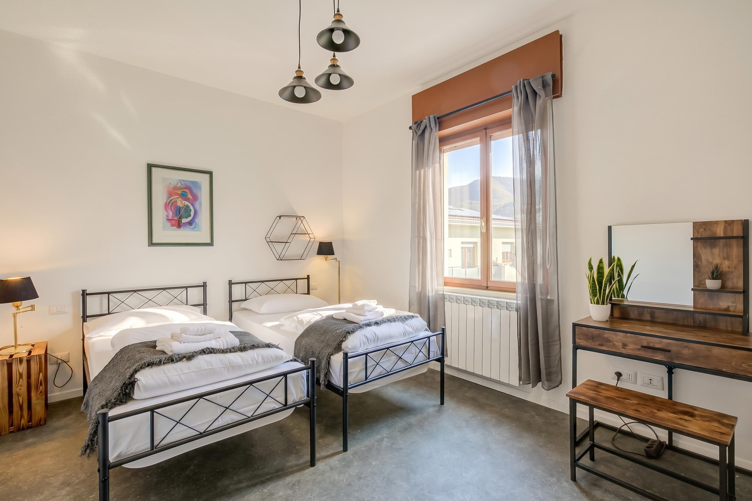 Een slaapkamer met twee bedden, een nachtkastje, een dressoir, een bankje, een raam met gordijnen en een spiegel aan de muur.