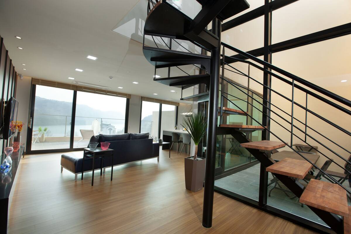 A modern living room with a black sofa, a coffee table, and a potted plant. There is a staircase with wooden steps and a glass railing.