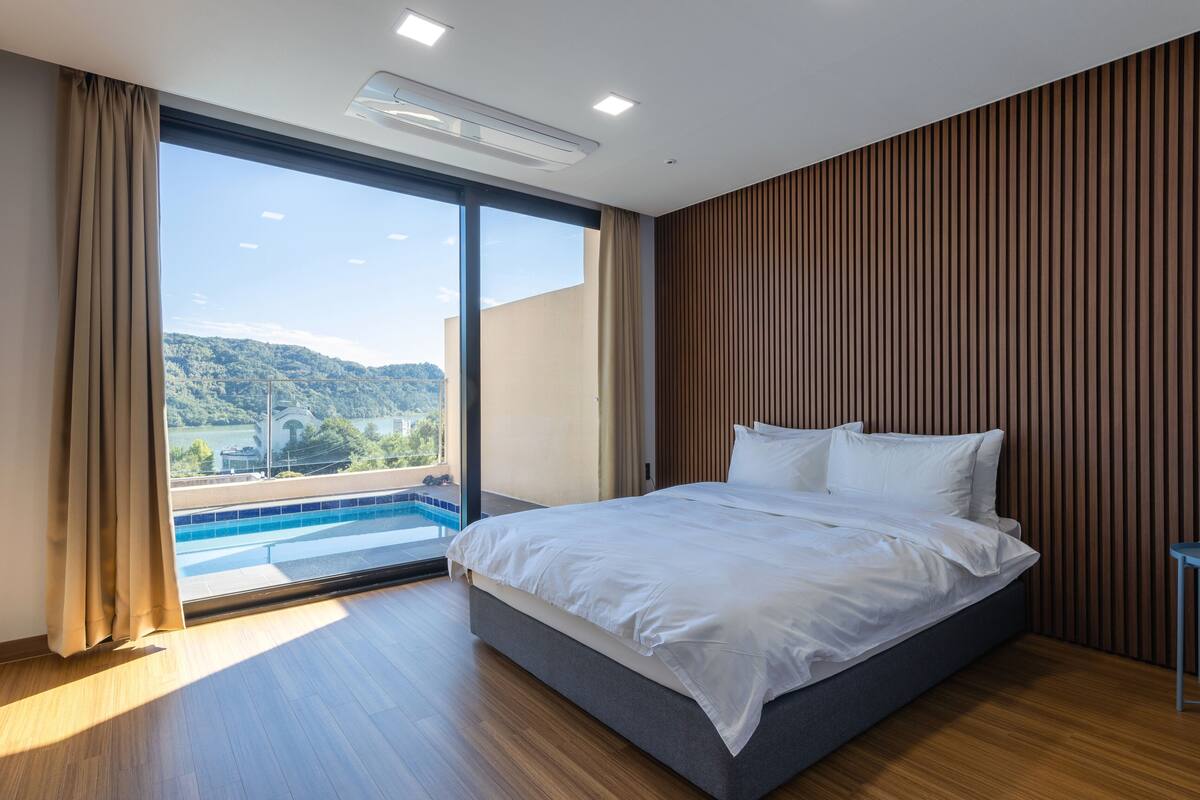 A modern hotel room with a large bed, a view of a swimming pool, and a wooden wall.