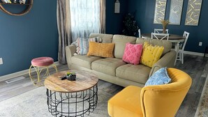 Een woonkamer met een beige bank, een gele fauteuil, een ronde salontafel en een eethoek met een witte tafel en stoelen.