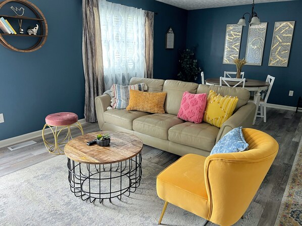 Un salon avec un canapé beige, un fauteuil jaune, une table basse ronde et un coin repas comprenant une table et des chaises blanches.