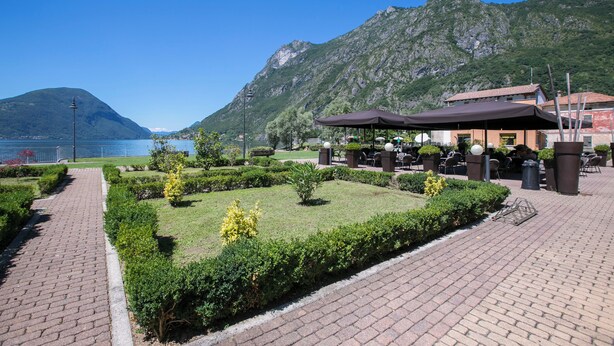 A paved walkway with a well-maintained garden and outdoor seating area, overlooking a body of water and mountains. - Porto Letizia Mozart 6, Porlezza, Italy (Porlezza)