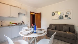 A modern kitchen with white cabinets, a dining table set with plates, cutlery, and bottles. A brown sofa and a painting are visible in the living area. - Porto Letizia Mozart 6, Porlezza, Italy (Porlezza)