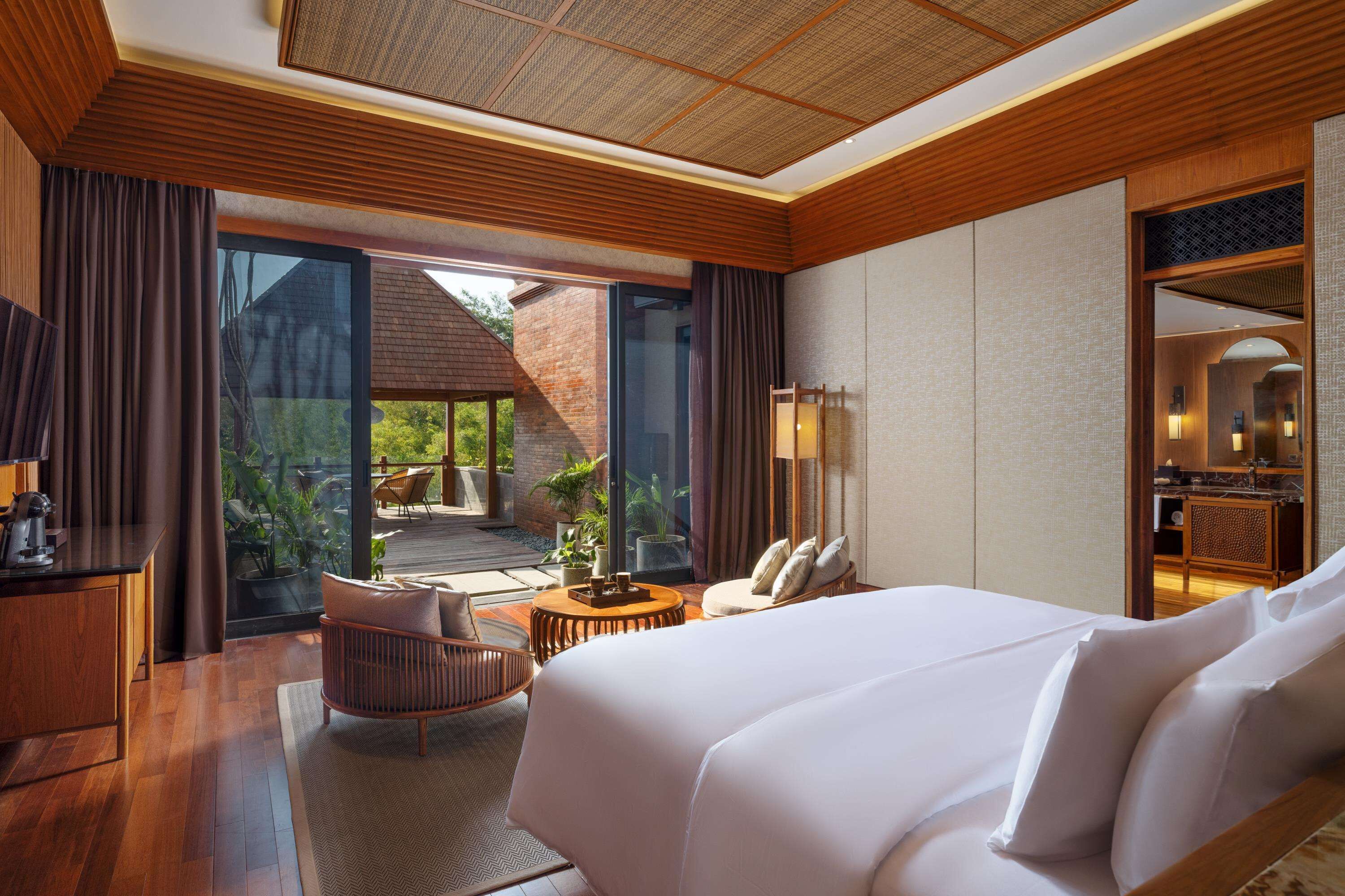 Habitación de hotel con una cama grande, un escritorio, una silla y vistas a un jardín por la puerta corrediza de vidrio.