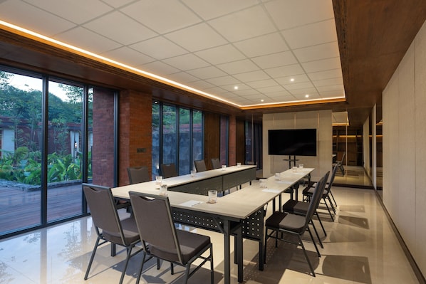 A conference room with a long table, chairs, a flat-screen TV, and large windows offering a view of greenery outside.