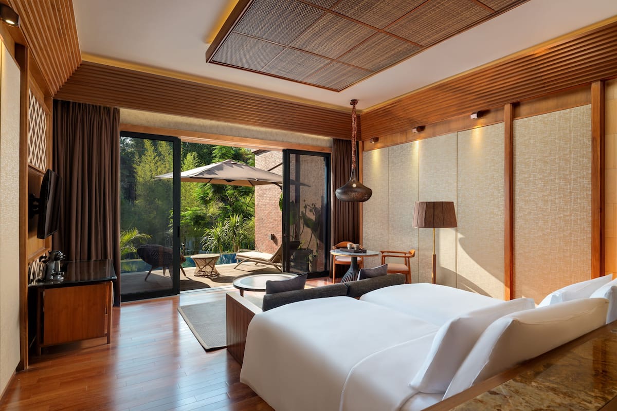 A modern hotel room with a large bed, a desk, a chair, a small table, and a view of greenery through the sliding glass door.