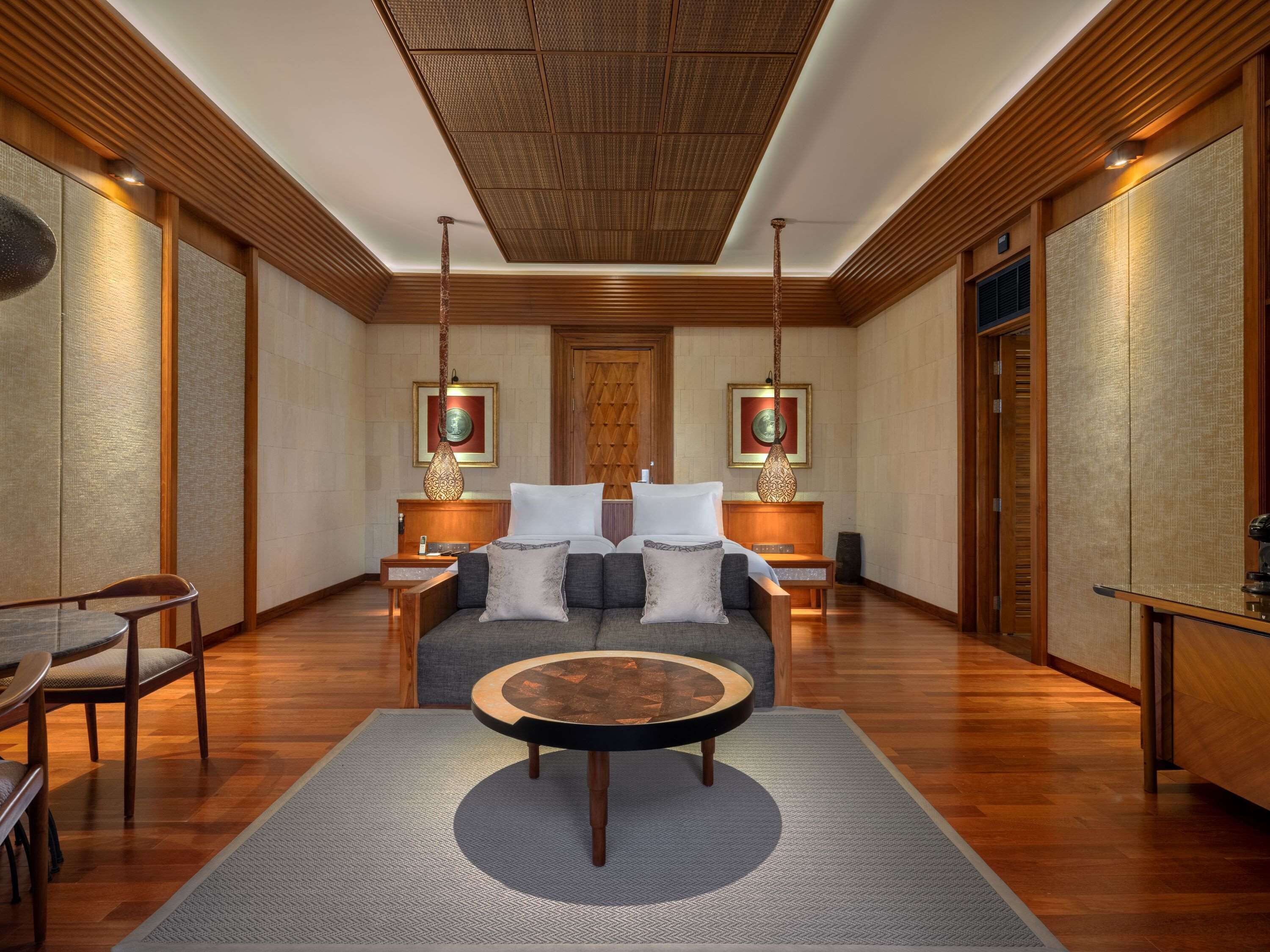 A modern hotel room with a central seating area, wooden paneling, and two wall-mounted lamps.