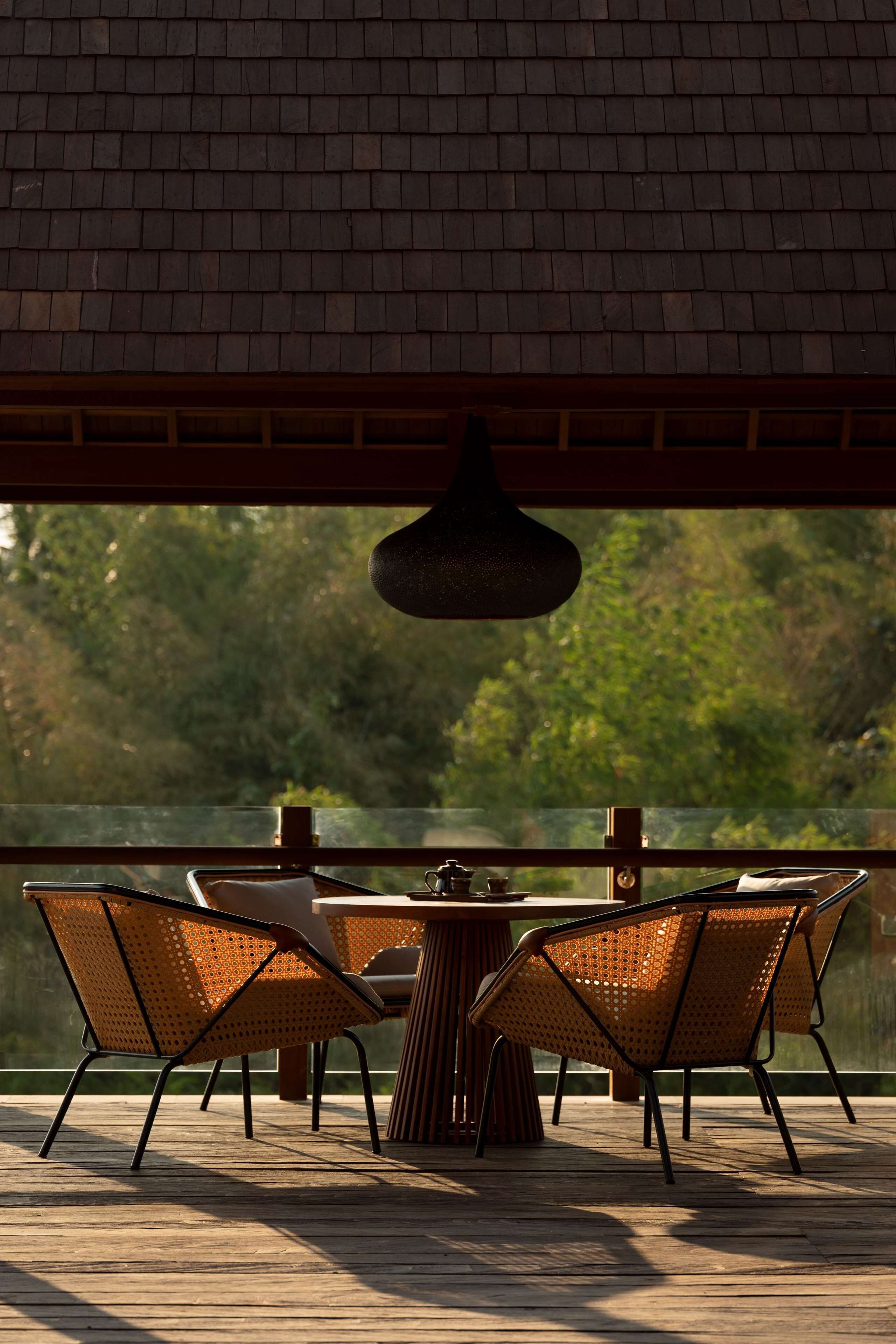 An outdoor patio with a round table and four wicker chairs, overlooking a body of water and greenery.