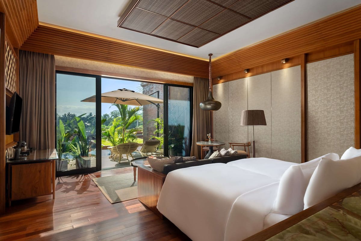 A modern hotel room with a large bed, a desk, a chair, and a view of greenery outside.