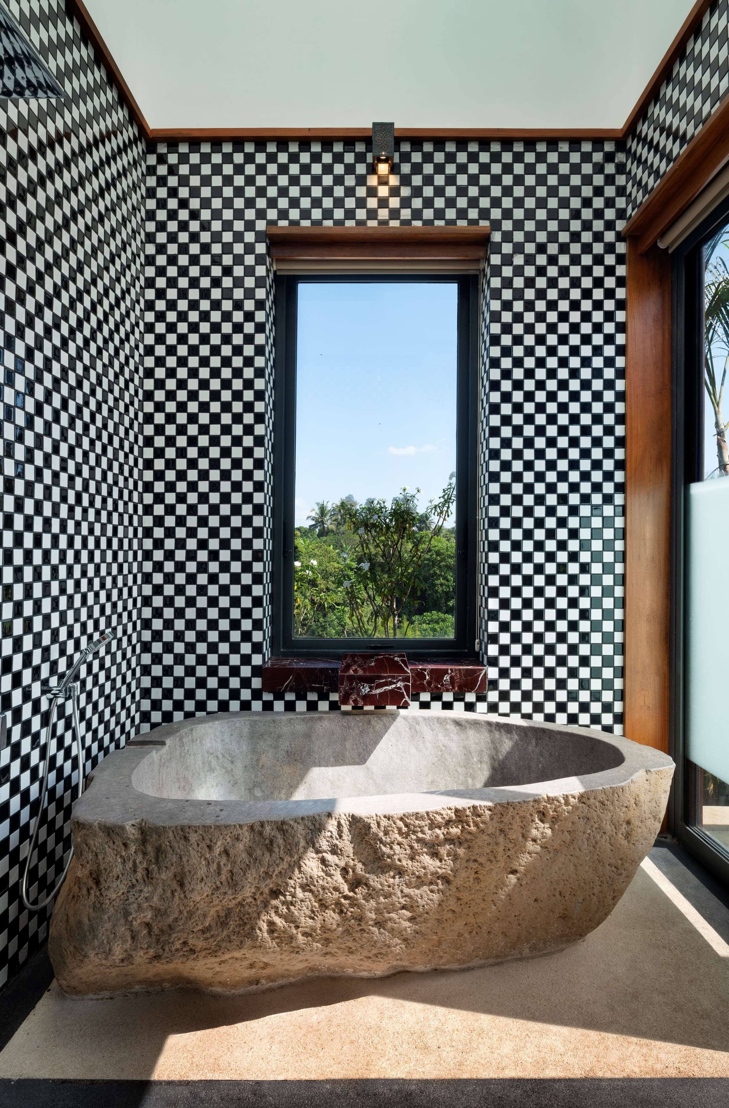 A modern bathroom with a large stone bathtub, checkered wall tiles, and a window offering a view of greenery.