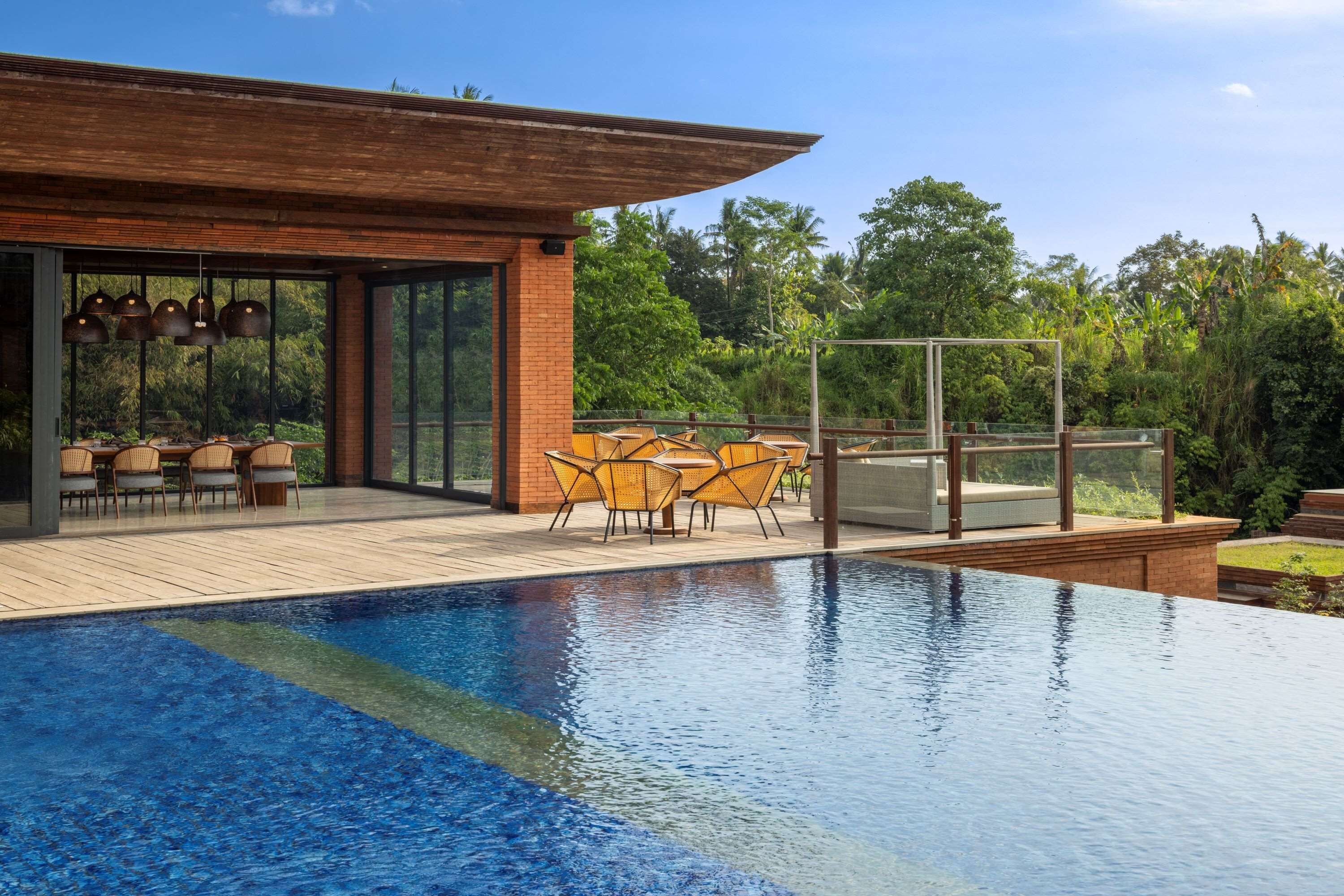 Un área de piscina moderna al aire libre con terraza de madera, mobiliario exterior y vistas a un follaje exuberante.