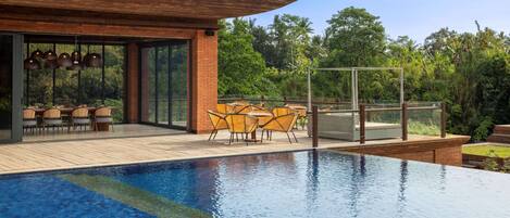 Un área de piscina moderna al aire libre con terraza de madera, mobiliario exterior y vistas a un follaje exuberante.