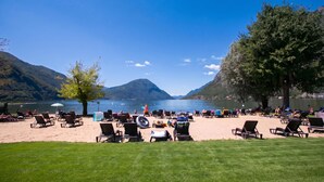 Ein Strand am See mit Liegestühlen und Tischen, umgeben von Bergen und blauem Himmel.