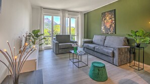 Uma sala de estar moderna com um sofá cinzento, um banquinho verde e uma mesa de centro. Há um quadro na parede e uma varanda com vista para as montanhas.