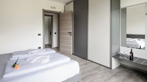 A modern hotel room with a neatly made bed, a bedside table with a bottle and glasses, and a large mirror reflecting the room.