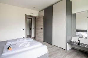 A modern hotel room with a neatly made bed, a bedside table with a bottle and glasses, and a large mirror reflecting the room.