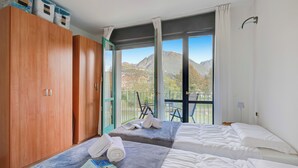 A hotel room with two beds, a wooden wardrobe, and a balcony with a mountain view. - Porto Letizia P5, Porlezza, Italy (Porlezza)