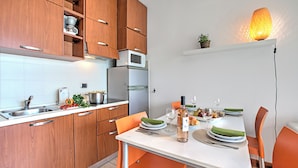 A modern kitchen with wooden cabinets, a white countertop, and a dining area with orange chairs and a white table. - Porto Letizia S11 B1, Porlezza, Italy (Porlezza)