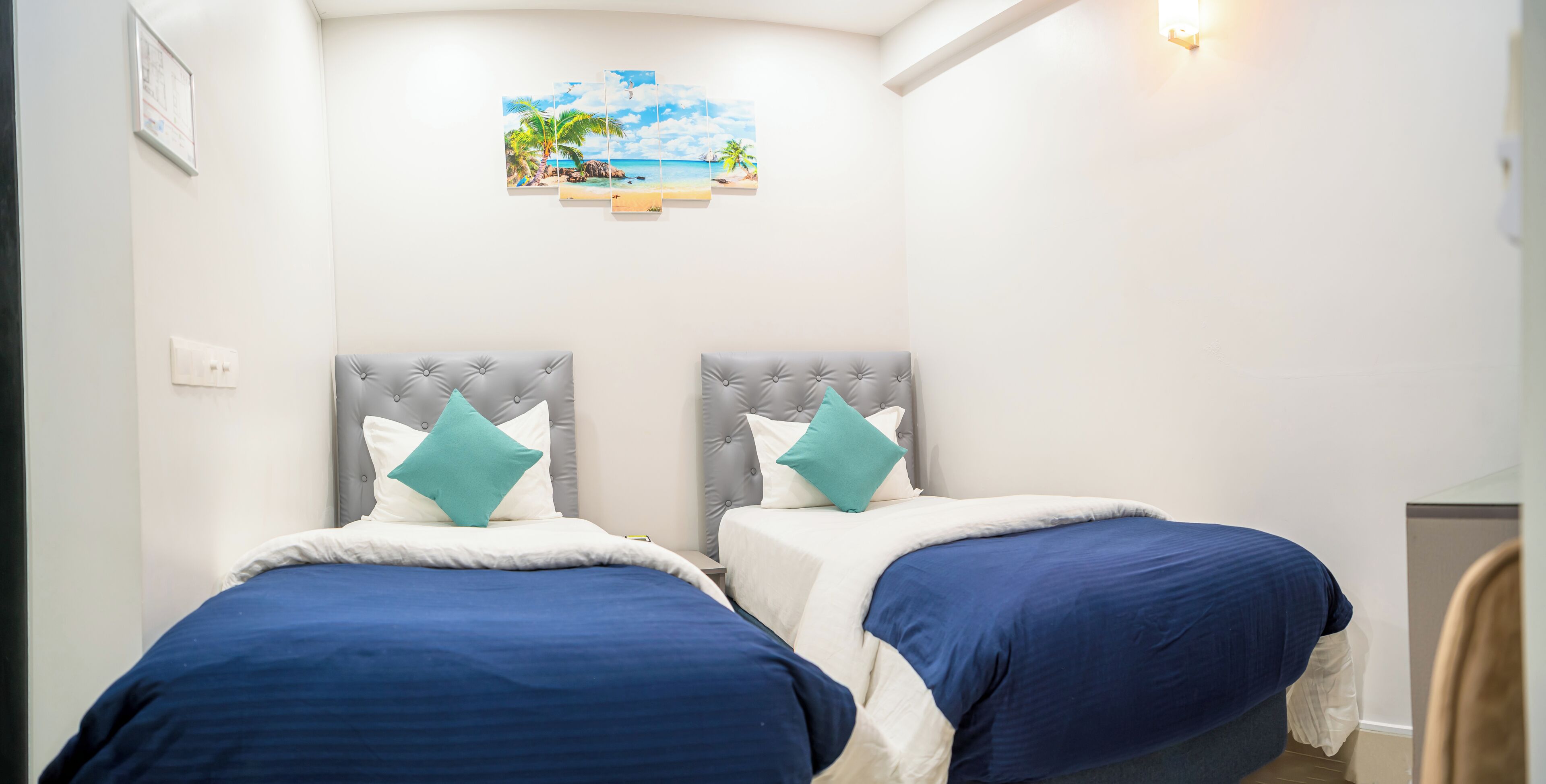 A hotel room with two beds, each with a tufted headboard and blue bedding. There is a wall-mounted artwork depicting a beach scene.