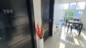 A modern room with a black door labeled '701', a white vase with flowers, a small table with chairs, and a view of buildings through large windows. - TOUR INN (Malé)