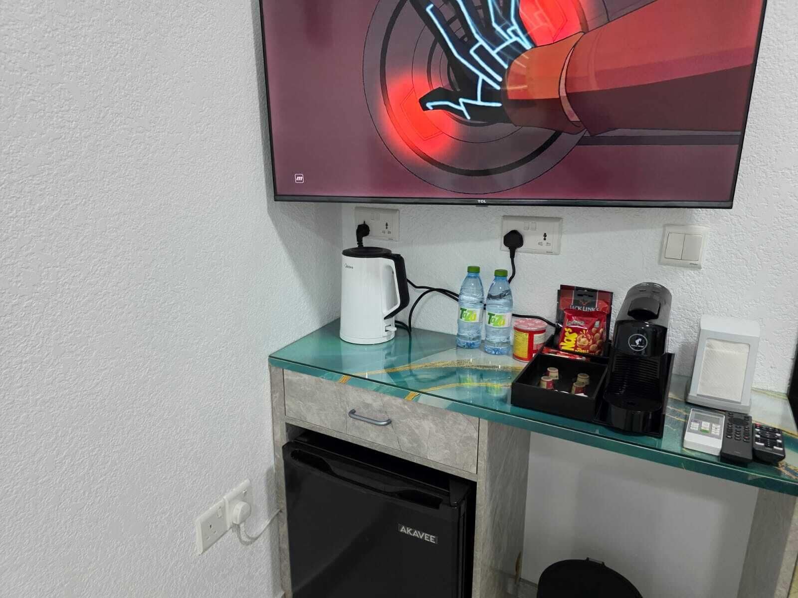 A hotel room with a glass coffee table, a white coffee maker, two water bottles, a snack box, a black coffee machine, a mini fridge, and a TV mounted on the wall.