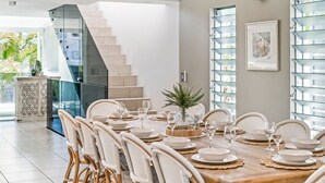 A dining area with a wooden table set for six, surrounded by wicker chairs. A staircase with glass railings is visible in the background.
