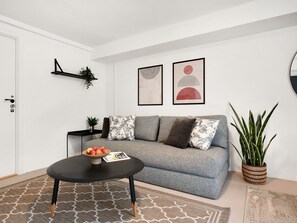 Ein Wohnzimmer mit grauem Sofa, einem schwarzen Couchtisch mit Fruchtschale, einer Zimmerpflanze und gerahmten Kunstwerken an der Wand.