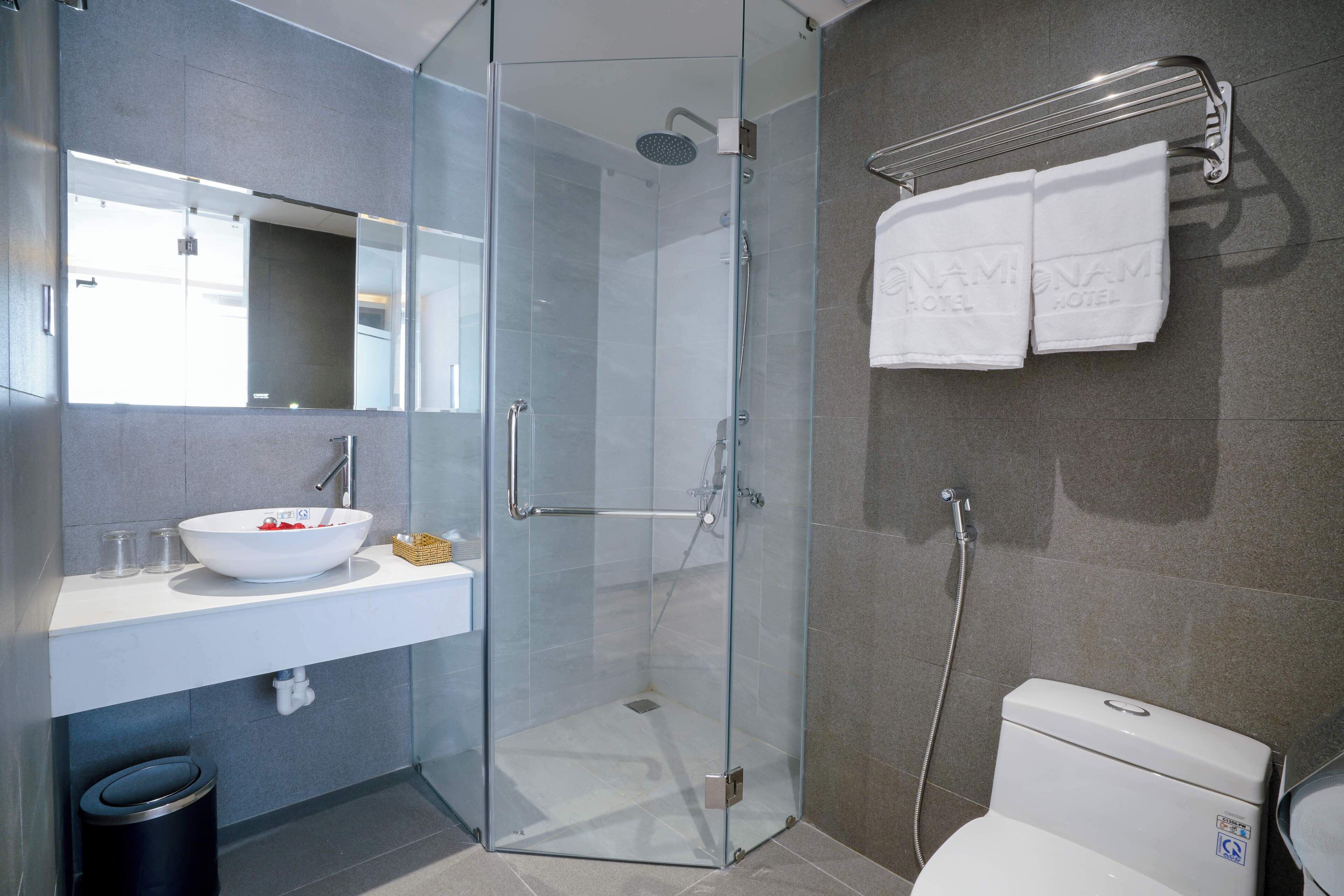 Un baño moderno con ducha envuelta en vidrio, lavabo blanco con un recipiente y un inodoro con una etiqueta visible.