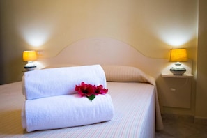 Una cama bien tendida con dos toallas y una flor decorativa, una lámpara de noche y una mesita de noche con un cuenco.