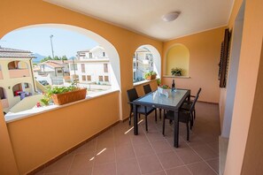 Un balcón con una mesa y sillas de vidrio, plantas en macetas y vistas a edificios cercanos.