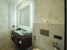 Un baño moderno con salpicadero de mármol, un espejo grande con iluminación integrada, un mueble de tocador oscuro, un perchero y un secador de manos.