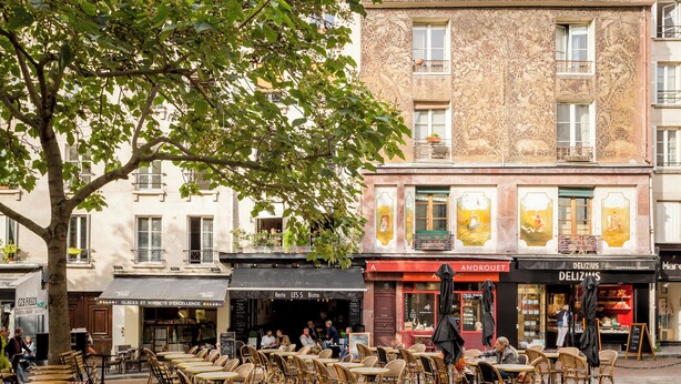 Un café al aire libre con mesas y sillas de mimbre, un árbol y edificios con escaparates al fondo.
