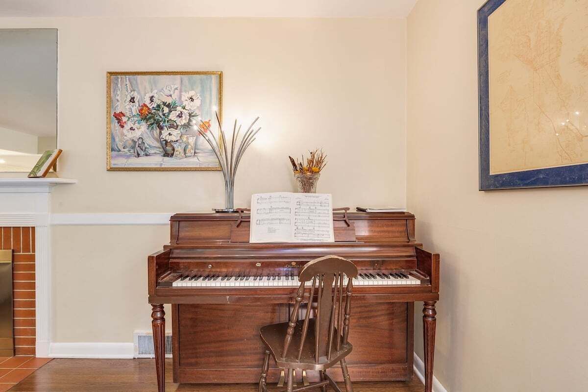 A piano with sheet music, a chair, a painting, and a vase with dried plants.
