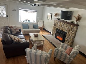 Ein Wohnzimmer mit Kamin aus Stein, einer Couch, zwei gestreiften Sesseln und einem Fernseher.