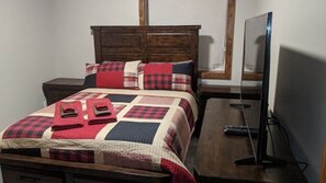 A bed with a quilt, a wooden headboard, a TV on a wooden stand, and a nightstand.