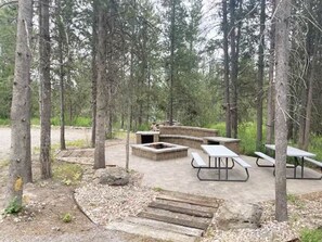 A forested campsite with picnic tables and a fire pit area.